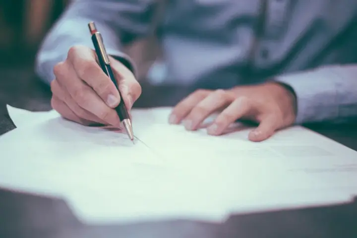 Mano firmando un contrato de reserva de vivienda. El documento se extiende sobre una mesa, con un bolígrafo en acción.