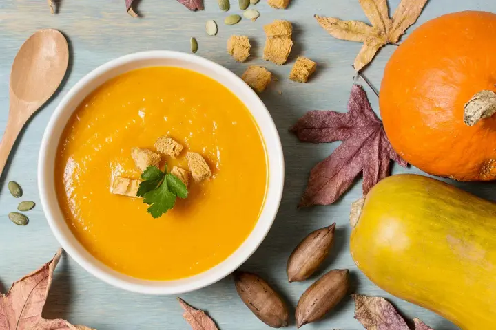 Un reconfortante tazón de puré de calabaza, adornado con picatostes y perejil, junto a calabazas, hojas de otoño y nueces.