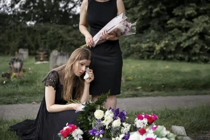 Smutna kobieta w czarnej sukience klęczy przy grobie, ocierając łzy. Obok stoi druga kobieta z bukietem kwiatów. To obraz żałoby, trudny urlop macierzyński po poronieniu.