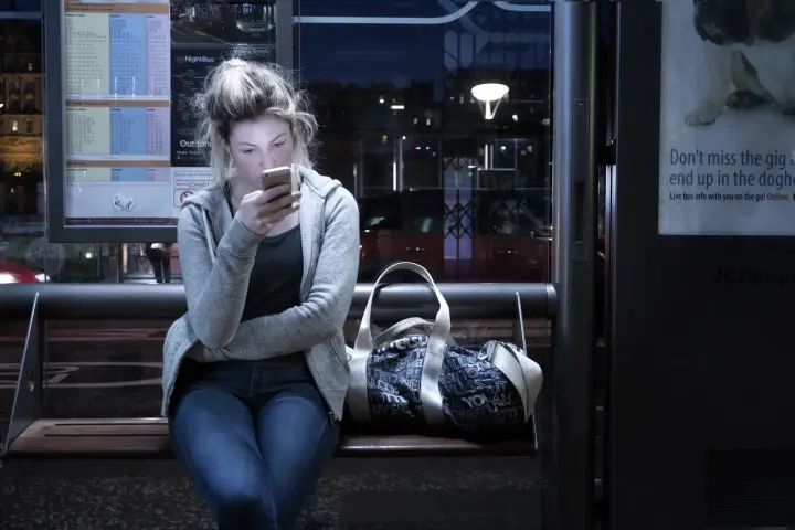 Dziewczyna na przystanku nocnego autobusu, pochłonięta telefonem, ignoruje uporczywe niepokojenie. Obok torba.