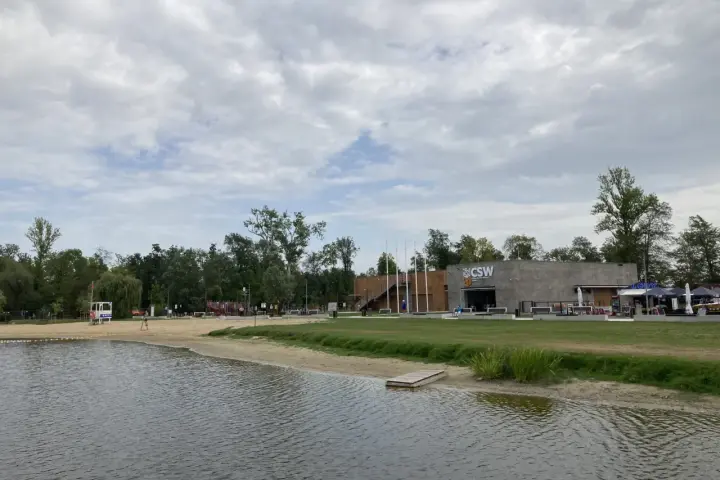 Spokojne jezioro małe żnińskie z piaszczystą plażą, budynkiem CSW i zielenią w tle.