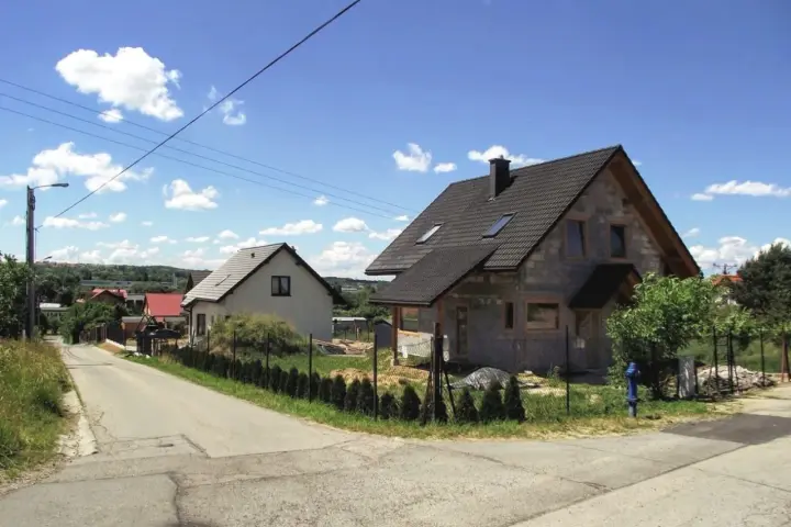 Dwa domy w budowie, jeden gotowy, na tle błękitnego nieba z chmurami. Widać drogę i zieleń. Różne kategorie gleby widoczne na poboczu.