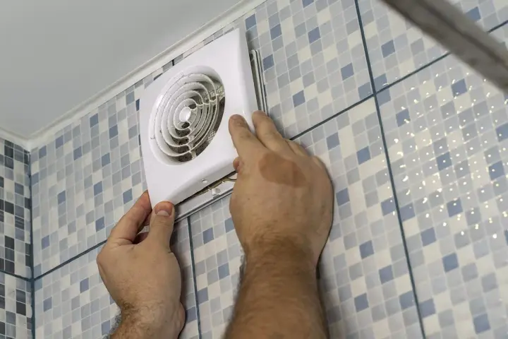 Mains d'un homme installant un ventilateur, une étape clé pour une bonne installation VMC dans une salle de bain carrelée.