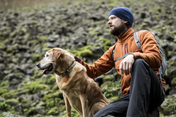 A man in an orange jacket and blue beanie rests during a hiking trip with his golden dog.