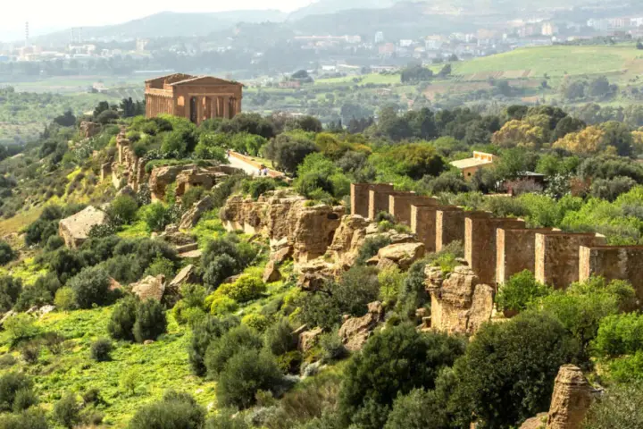 Ancient Greek temples stand proudly on a ridge in the Valley of the Temples, Sicily, surrounded by lush greenery and a distant town.