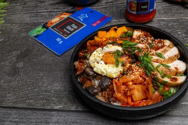 Pyszny Bibimbap przepis: ryż, kurczak, jajko sadzone, grzyby, marchewka i kimchi. Obok opakowanie pasty kimchi.