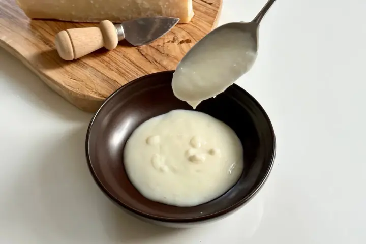 Un cucchiaio versa una cremosa crema di parmigiano in una ciotola scura. Sul tagliere, un pezzo di Parmigiano Reggiano e un coltellino.