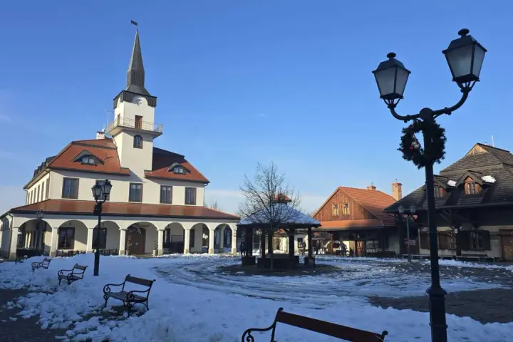 Zimowe miasteczko galicyjskie z ratuszem i karczmą, plac pokryty śniegiem, ławki i latarnie ozdobione świąteczną girlandą.