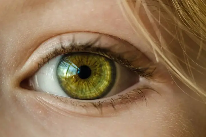 Zielona soczewka oka z refleksami światła, otoczona jasną skórą i blond włosami.
