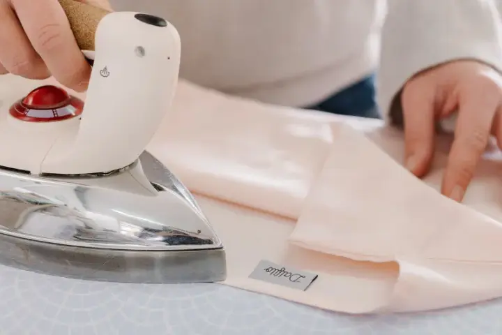Mani che stirano lenzuola rosa pallido con un ferro da stiro Immetec. Un'etichetta con il logo "Daffodil" è visibile sul tessuto.