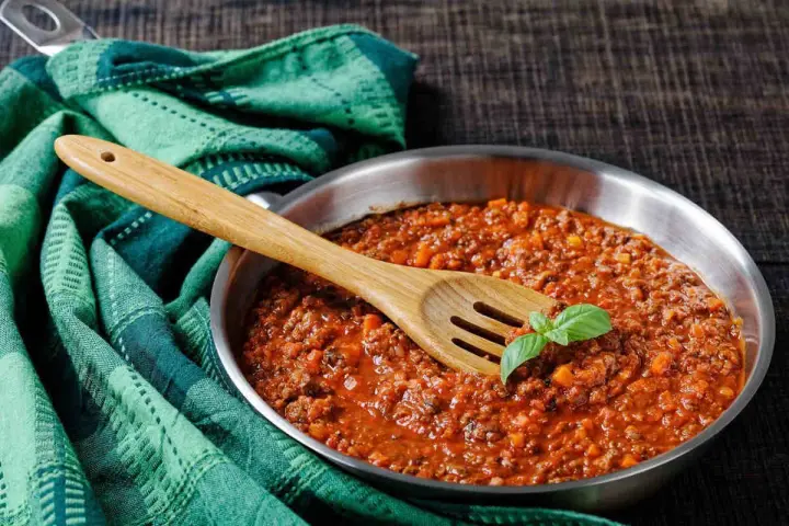 Un saporito ragù di cinghiale in padella, pronto per condire la pasta. Un cucchiaio di legno e una fogliolina di basilico completano la scena.