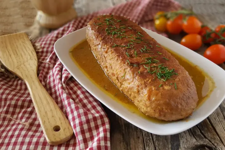 Polpettone in padella dorato, guarnito con erba cipollina, servito su un piatto bianco con salsa. Accanto, una spatola di legno e pomodorini.