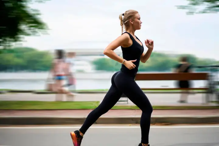 Donna bionda in abbigliamento sportivo nero corre con una postura corsa perfetta lungo un percorso alberato.