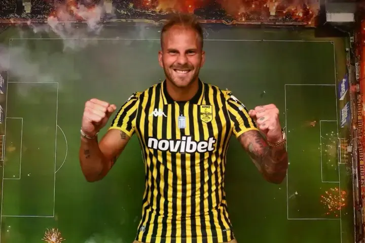 Uros Racic, con su nueva camiseta a rayas amarillas y negras, celebra con los puños en alto. El fondo simula un campo de fútbol con bengalas.