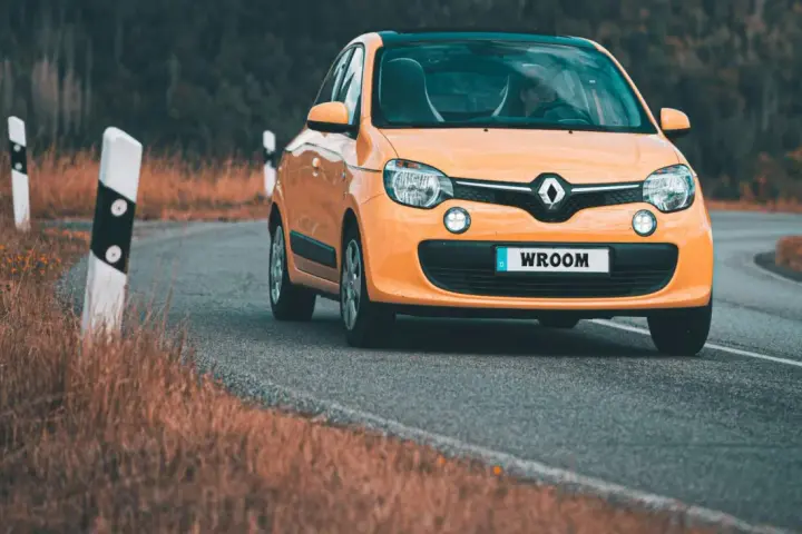 Une Twingo orange roule sur une route sinueuse bordée d'herbes sèches.