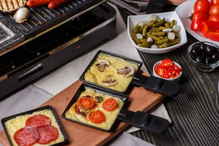 Raclette z pieczarkami i pomidorami, obok kiełbaski. Idealny ser do raclette rozpuszcza się w tych małych patelenkach.