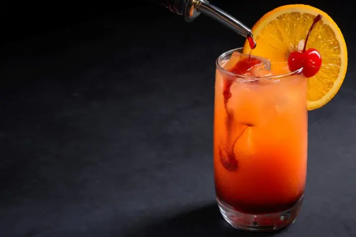 Kolorowy drink z syropem grenadina, ozdobiony plasterkiem pomarańczy i wisienką.