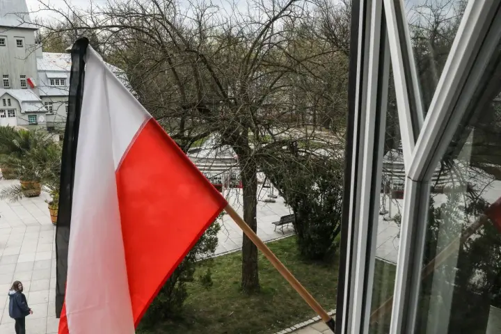 Flaga państwowa w biało-czerwonych barwach powiewa na wietrze. W tle widać drzewa i budynki.
