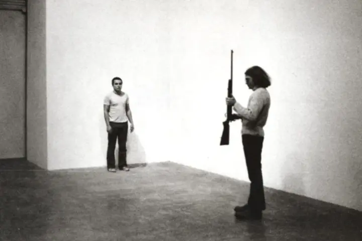 Chris Burden, dans une performance tendue, tient un fusil. Un homme se tient à distance, le regard fixé.