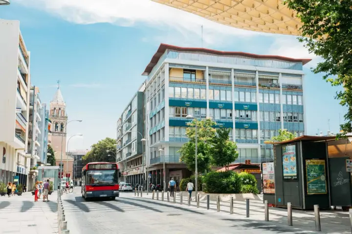 Un bus rouge circule dans une rue bordée d'immeubles et d'un clocher, sous un ciel bleu.