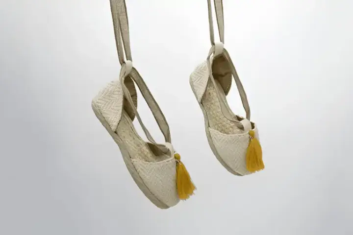 Dos alpargatas de tela beige con pompones amarillos cuelgan de tiras, mostrando su diseño veraniego.