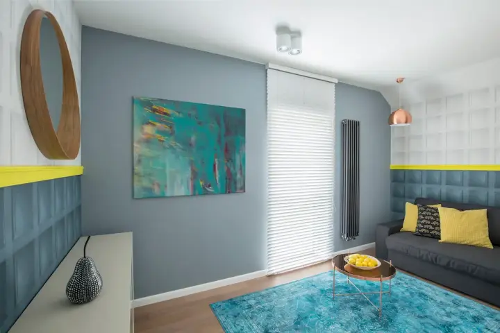 Sala de estar con sofá gris, alfombra turquesa y arte abstracto en tonos fríos. Un espejo redondo de madera y detalles en amarillo complementan el espacio.