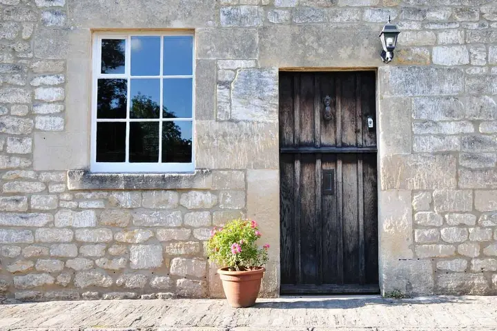 Facciate in pietra e mattoni grezzi con una porta in legno scuro e una finestra bianca. Un vaso di fiori rosa aggiunge un tocco di colore.