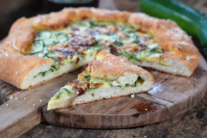 Leckeres Zucchini Pizza Rezept auf einem Holzbrett, mit einem Stück abgetrennt.