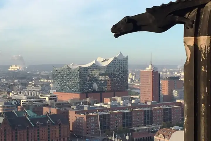 Blick über Hamburg mit der Elbphilharmonie, gesehen durch ein Fenster des Mahnmals St. Nikolai. Ein Wasserspeier wacht über die Stadt.
