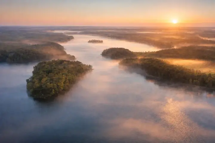Mglisty poranek nad jeziorem Mukrza. Słońce wschodzi nad lasami, tworząc złote refleksy na wodzie i unosząc się nad mgłą.
