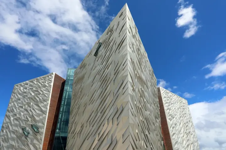 Nowoczesna bryła Muzeum Titanica w Belfaście, z geometrycznymi panelami i szklanymi elementami, pod błękitnym niebem.
