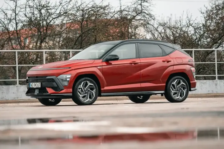 Czerwony Hyundai Kona Hybrid z czarnym dachem stoi na mokrym parkingu. Elegancki design i nowoczesne linie podkreślają jego hybrydowy charakter.
