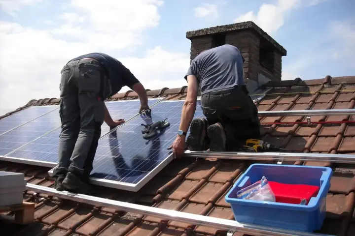 Dwóch monterów na dachu układa panele do instalacji PV. W tle komin i błękitne niebo.