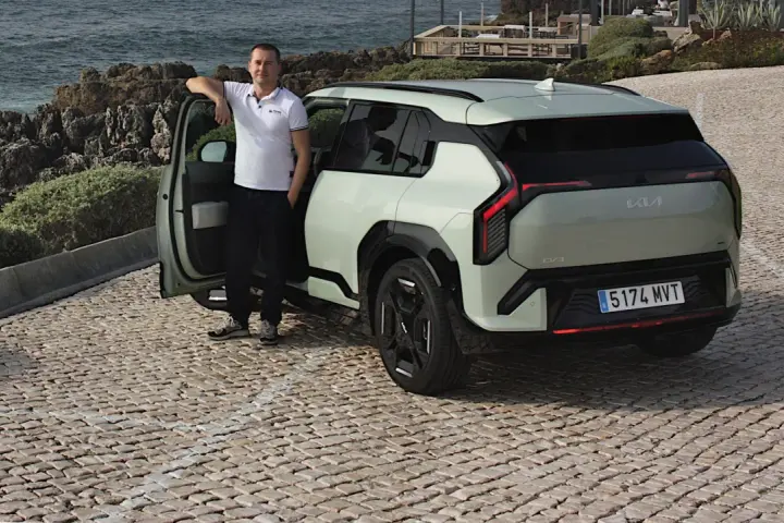 Mężczyzna opiera się o drzwi nowego, jasnoszarego Kia EV3, zaparkowanego na brukowanej drodze nad morzem.