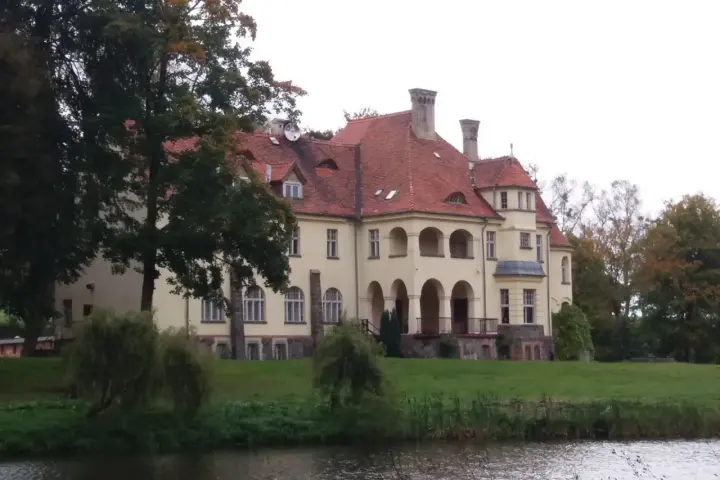 Malowniczy pałac z czerwoną, dachówkową, spadzistą dachem, otoczony zielenią i wodą.