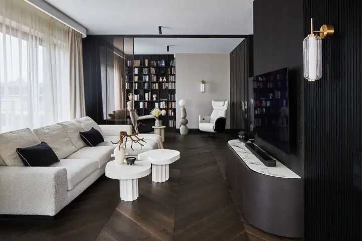 Nowoczesny salon z wygodną sofą, stolikami i regałem pełnym książek. Dominują ciemne, grafitowe akcenty, tworząc elegancką atmosferę.