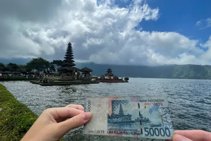 Ręka trzyma banknot 50 000 rupii na tle świątyni na wodzie. Czy Indonezja jest bezpieczna? Tak, podróżowanie po niej jest bezpieczne.