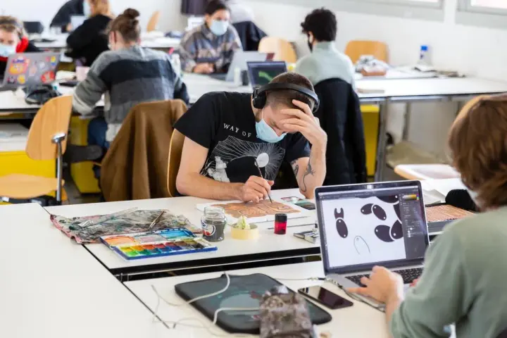 Des étudiants concentrés dans une école d'art, certains peignent, d'autres travaillent sur ordinateur. L'ambiance est studieuse.