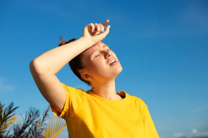 Une jeune femme en t-shirt jaune, le visage marqué par la chaleur, essuie son front sous un ciel bleu intense. La durée d'une insolation est palpable.