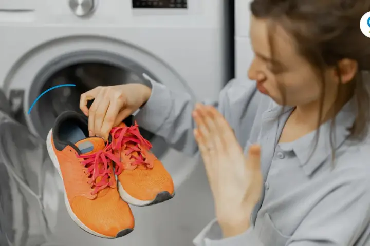 Woman holding smelly orange sneakers near an open washing machine.
