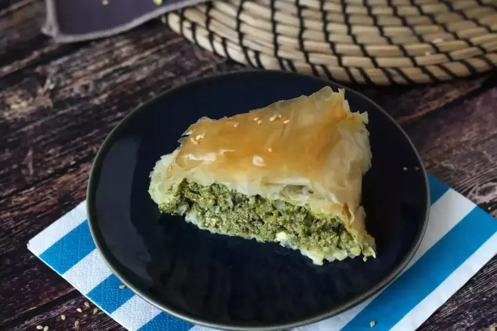 Kawałek spanakopity z chrupiącym ciastem filo i zielonym nadzieniem ze szpinaku i sera feta, posypany sezamem.