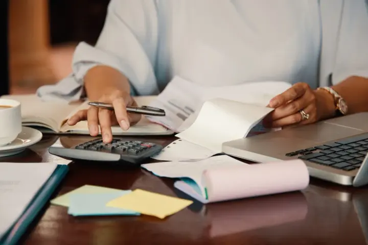Persona revisa documentos y usa calculadora para el modelo 184. Hay café, portátil y notas adhesivas.