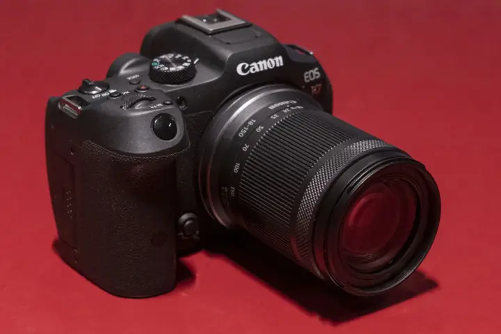 Appareil photo noir Canon EOS R7 avec objectif 18-150 mm, posé sur un fond rouge vif.