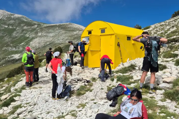 Gruppo di escursionisti vicino a un giallo blockhaus Majella, su un sentiero roccioso con montagne verdi sullo sfondo.