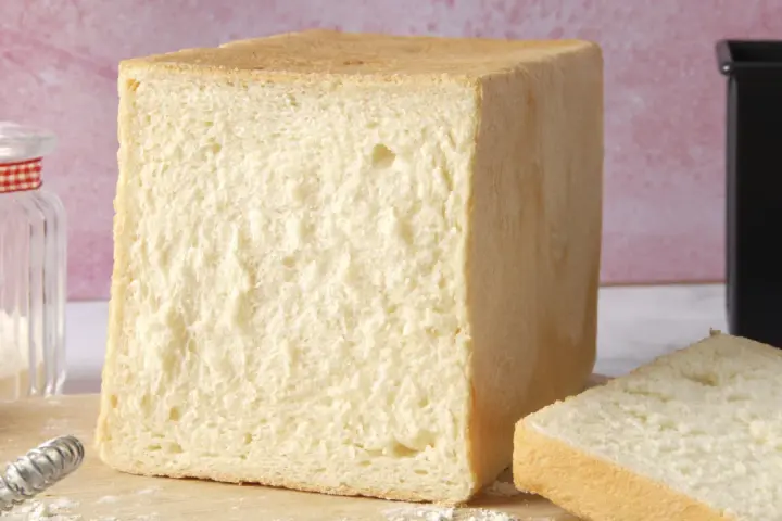 Un pain de mie moelleux, parfait pour une recette facile. Un pot de farine et un moule à pain sont à proximité.