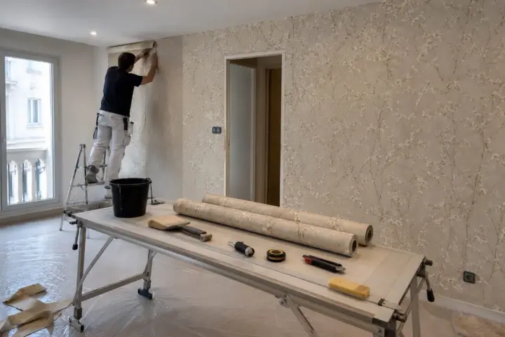 Hombre en escalera empapelando pared con diseño floral. Sobre la mesa, rollos de papel, brochas y herramientas para empapelar.