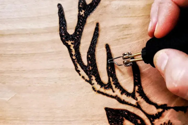 Dettaglio di un disegno a pirografia su legno: una mano guida le punte pirografo per creare le corna di un cervo.