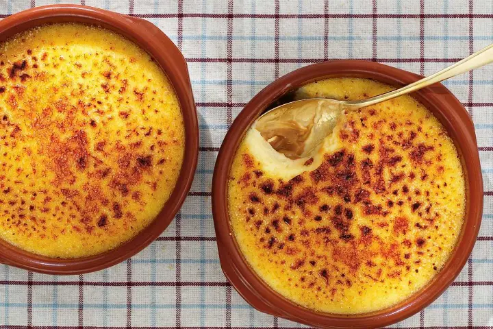 Två krämiga creme brulee i bruna skålar, en med en gyllene sked som lyfter fram den karamelliserade ytan.