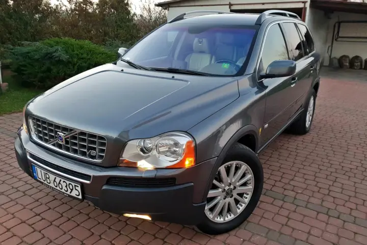 Szary Volvo XC90 z silnikiem V8, zaparkowany na podjeździe z kostki brukowej, otoczony zielenią.