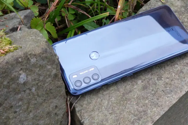 Błyszczący tył telefonu **Moto G 50** z potrójnym aparatem, leżący na kamieniach wśród zieleni.